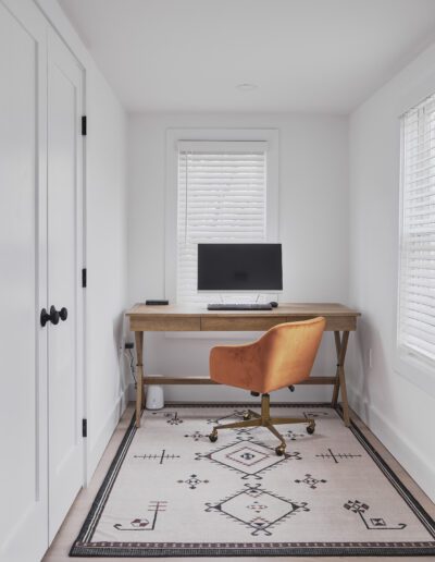 A minimalist home office with a wooden desk, an orange office chair, a computer monitor, and a patterned rug. The room features white walls, a window with blinds, and a closet with double doors.