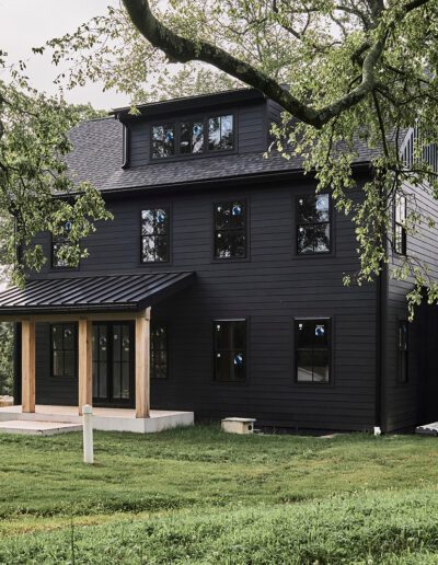 A modern black house with large windows sits on a grassy lawn, with a tire swing hanging from a tree in the foreground and construction materials on the right side.