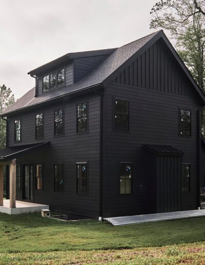 A modern black two-story house with multiple windows, a covered front porch with wooden posts, and a grassy yard surrounded by trees.