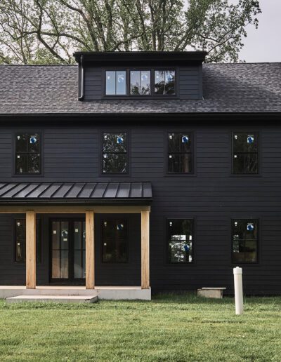 A modern two-story house with black siding, large windows, a covered front porch with wooden columns, and a grassy yard surrounded by trees.