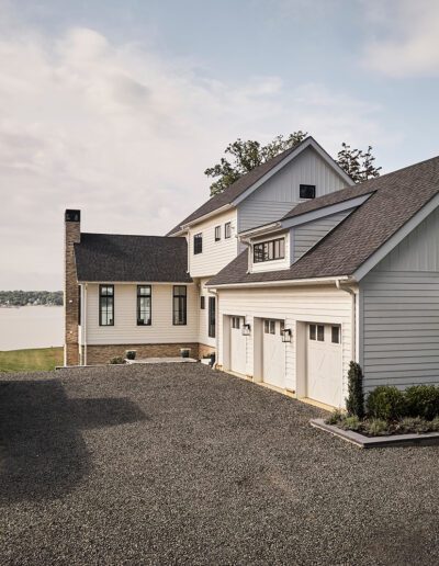 Two white detached houses with dark roofs are shown beside a gravel driveway, with a view of water and trees in the background on a partly cloudy day.