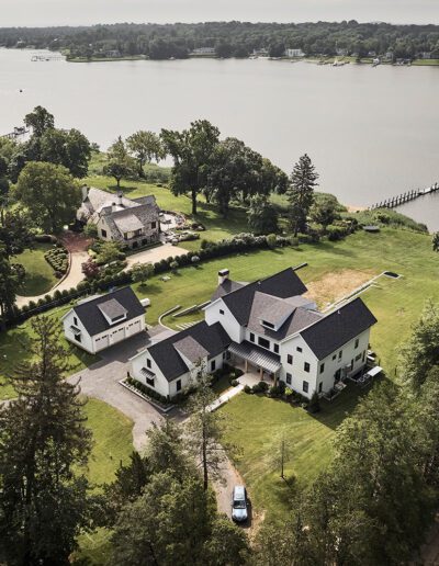 Aerial view of a large white house with several surrounding buildings, a driveway, and a green lawn, located near a river with trees and other houses nearby.