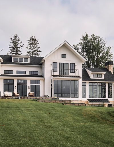 A large modern farmhouse-style home with white siding, black-framed windows, and multiple gabled roofs, set on a green lawn with trees in the background.