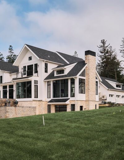 Large modern farmhouse with white siding, black-framed windows, stone chimney, and multiple levels, surrounded by green lawn and trees; detached garage visible in background.