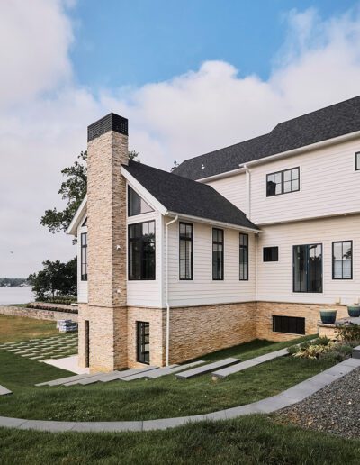 Modern two-story house with light siding and brick details, featuring large windows, a sloped yard, and a view of a lake under a partly cloudy sky.