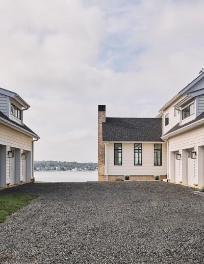 Two white, modern farmhouse-style buildings with gable roofs frame a gravel driveway, leading to a house overlooking a lake under a cloudy sky.