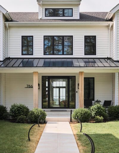 White two-story modern farmhouse with black-framed windows, a metal roof over the porch, wooden pillars, landscaped front yard, and a paved walkway leading to the entrance.