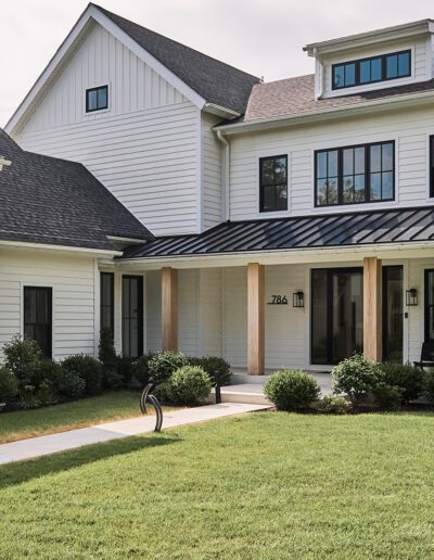 A modern two-story white house with multiple gables, a covered porch with wooden columns, and a well-kept lawn with shrubs and a curved walkway.