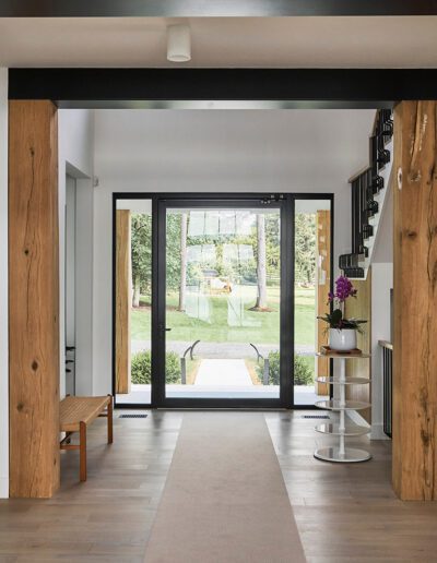 Modern hallway with exposed wooden beams, light wood flooring, a bench, potted plants, and a large glass door leading to a green backyard.
