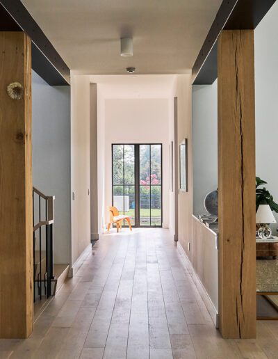 A modern hallway with light wood floors, wooden pillars, black metal stair railings, and a glass door at the end leading to a garden with trees outside.