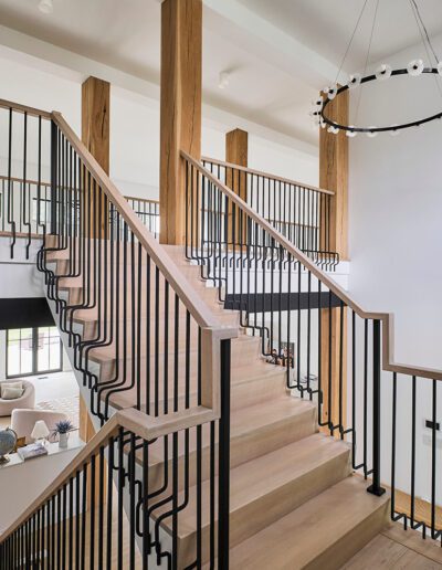 A modern wooden staircase with black metal railings leads to an upper floor; large vertical wooden posts and a circular chandelier are visible.