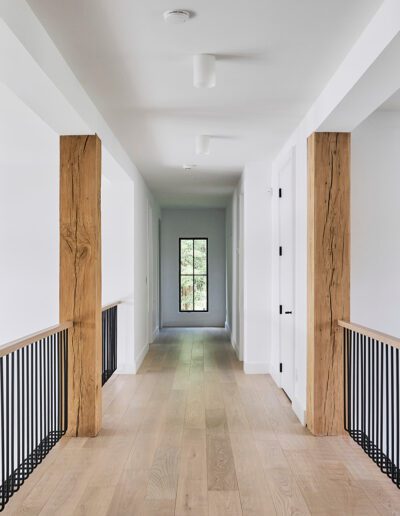 A bright hallway with wooden floors, exposed wooden beams, white walls, black metal railings, and a glass door at the far end.